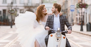 Het bruidspaar rijdt op een scooter door een straat in de stad, glimlachend en samen genietend van een romantisch huwelijksmoment.